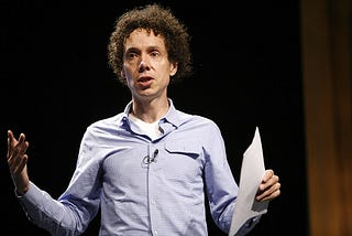 Malcolm Gladwell giving a lecture, holding a piece of paper in his hand