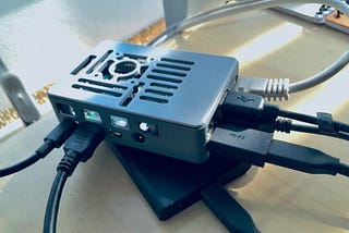 Photograph of a Raspberry Pi computer. It’s in a grey plastic housing and underneath is a SSD drive. Wires are sticking out from it on all sides.