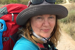 woman wearing sun hat, sunglasses, red backpack, walking a long trail