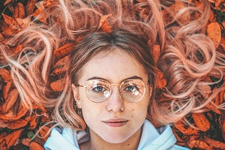 A young woman lying on a bed of fallen leaves. The leaves are red. The woman’s long hair is brown, dyed pinkish-blonde at the ends, and is spread out around her head. She is wearing a pale blue hoodie and wire-framed glasses with rounded lenses. She has a slight, enigmatic smile.