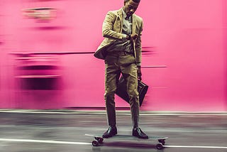 Professional Black man wearing tan suit, carrying briefcase and riding a skateboard down a street while looking at his watch.