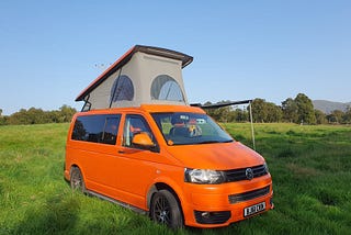 orange T5 campervan with a pop top up parked on grass with blue sky above it