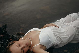 A woman in a white dress lies peacefully at the edge of dark water, eyes closed, symbolizing stillness, reflection, and emotional healing.