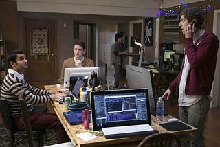 A screenshot from HBO’s “Silicon Valley” television show where three middle-aged men are gathered around a rectangular table full of computers (two people are seated, one is standing on his phone). The table is messy and filled with water bottles, disposable coffee containers, and office knick knacks.