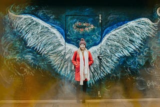 woman in red coat standing in front of large angel wings and crown of flowers painted on a wall.