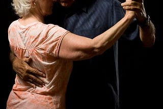 An older couple hold one another in old fashioned slow-dance style, one hand on the other’s back, their other hands outstretched and interlocked.