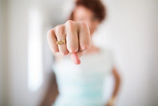 I woman stands with her fist in the camera. The fist is in focus and the woman is out of focus in the background. The woman wears a gold ring on her ring finger that says “I am bad ass”