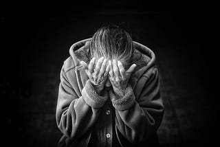 Black and white image of person wearing a sweatshirt holding their head in their hands. Their hair is long and tied back.