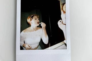 A polaroid of the author in her wedding dress, looking into a mirror and applying red lipstick