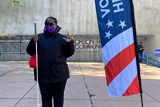 Casandra standing near a “Vote Here” flag. Her face is masked. She has a cane in right hand and left hand showing ILY sign.