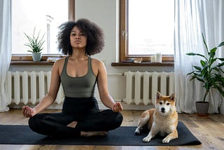 A young woman sits in a lotus pose on a yoga mat with her dog laying next to her.