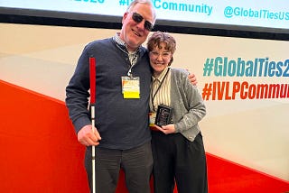Two World Learning staff members (a man and a woman) stand side by side in front of a large backdrop in red and white. The man holds a walking cane, and the woman holds an award plaque.