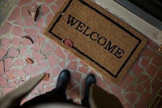 Photo looking down from the perspective of a person standing on the edge of a brown Welcome mat, on top of brick tile, with Autumn leaves scattered around, like a snapshot of idealized White suburban home entrances.