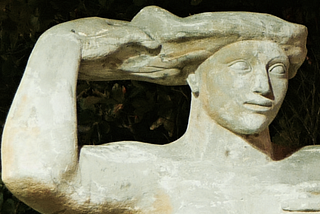 A close up view of the head and arms of a large statue. The statue’s face is tranquil, and the lines are drawn as in Ancient Egyptian statues. With one arm the figure reaches up to touch her hair, which unfurls in a way which suggests the surface of the sea.