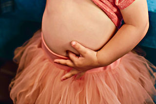 Picture of a toddler girl wearing a peach-colored tutu and top. Her top is pulled up, and her left index finger sticking into her navel.