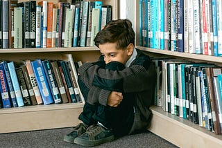 A young boy sits curled up in the corner of a library, hugging his knees to his chest with a worried or anxious expression.