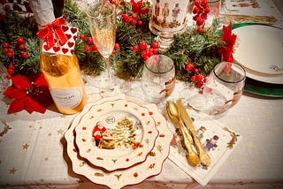 A table laid out for a Christmas meal, complete with seasonal plates, sparkling champagne flutes, and plastic gold cutlery. There are also pine and holly garlands, poinsettias, a candle featuring nutcrackers, and a gold champagne bottle of chocolate con leche. The main place setting has dishes that feature a scene with Santa Claus, and there are some glasses with nutcrachers, as well. There is a tablecloth with gold stars on it.