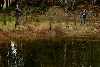 Two people stand about 15 metres apart on the edge of a pond arms crossed, backs stiff, frowning at each other