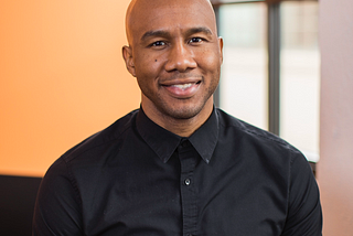 Javier, a bald Black man, wears a black button down shirt and is smiling with teeth visible.