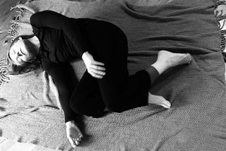 B/w photo of dancer lying on her side on a blanket, dressed in black, head resting on the floor, arms loose, legs bent.