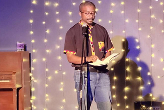 Photo of Professor Dr. Doug Manuel reading from his newest poetry collection “Trouble Funk” in front of lights.