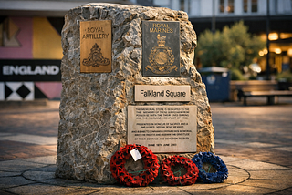 A granite memorial stone in Falkland Square, Poole, UK, displays plaques for the Royal Artillery and Royal Marines, with a dedication to local servicemen who died in the Falklands conflict. Two wreaths of red and blue poppies rest at its base. Warm evening light highlights the stone’s texture while the square and shops blur softly in the background, evoking quiet remembrance amid everyday life.