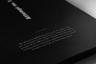 A photograph of the cover of the short visual book I designed for this project. The photo is taken from an angle in front of it sitting on my desk and is a black and white photo. The design of the book is minimal with the title of the book (“attempt no. 3") in a white, bold font with the word rotated couner clockwise in the centre of the book cover, so you read the title from the bottom up. Below the title is what looks like a short description of the book but is not legible.