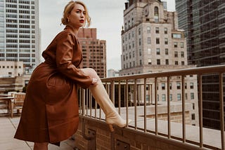 A blonde woman of indeterminate age, wearing red lipstick, is standing with one foot raised and resting on a low wall, so she can adjust her knee-high, stack-heeled, white leather boot. She is wearing a knee-length trench coat, and no hint of a skirt can be seen on the thigh of her raised leg.