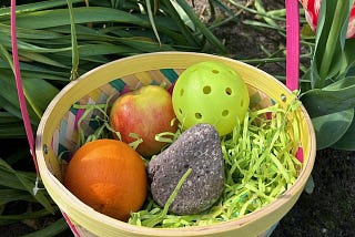 Photo of an Easter basket in an outdoor setting with a plastic wiffle ball, apple, organge, and rock in a bed of fake Easter grass.
