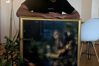 An artist sitting in a room and holding a framed painting.