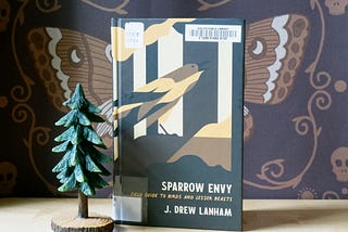 A book is propped on a wooden table. The book’s cover depicts a geometric image of a bird. Next to the book is a wooden figurine of a pine tree. In the background, a partially-obscured wall hanging features a stylized illustration of a death’s-head moth by graphic artist Lathe and Quill.