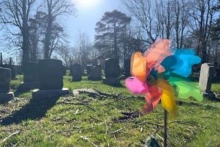 A rainbow colored pinwheel, softly spinning in the foreground. Behind it are many gravestones.