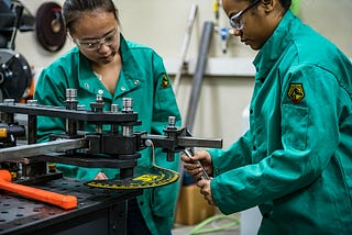 Two young women in teal safety jumpsuits work together in a machine shop.