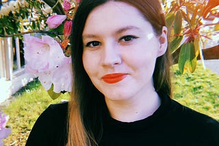 A photograph of Jenna James, a woman standing in front of a cherry blossom tree, wearing red lipstick and a black sweater.