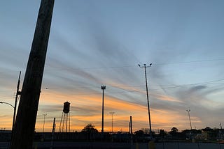 A sunset of pink and grey at the horizon in a light blue sky. a shadow of a water tower, electrical poles / wires, & a street