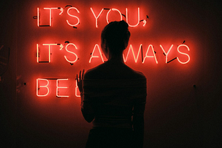 A backlit young woman standing in front of a red neon sign that reads “it’s you, it’s always been…”