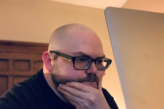 A bald person with glasses and a beard angsts looking at a laptop screen.