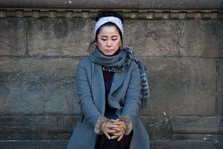 A woman sits quietly next to a stone wall with hands folded and eyes downcast