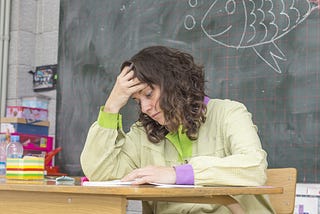 A white woman with brown hair sits at her desk in a classroom in front of a blackboard. She has her head resting in her hand, and her eyes stare blankly down at a paper.