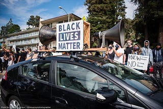 A car with two large bullhorns and a large sign that says “Black Lives Matter” on the root in the midst of a protest crowd.