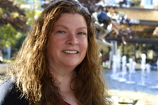 Photo of author JoEllen Claypool with shoulder-length brown hair with a burgundy shirt and black cover shirt with fountains in the background.