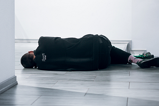 [Image: Person in black clothes lying on gray floor in room with white walls. They are facing away from the viewer.]