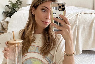 Tara Michelle sitting on the floor in front of her bed, holding a coffee. She is taking a mirror selfie with her phone.