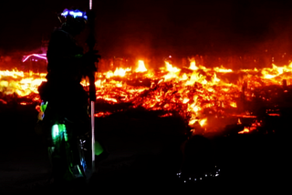 A nighttime scene with an expansive fire casting an eerie glow. A barely visible female figure kneels on the desert ground, facing the flames. Her back, adorned with fairy lights, is exposed. To her left, a tall person with neon-glowing accessories holds a staff, watching her as if witnessing something sacred.