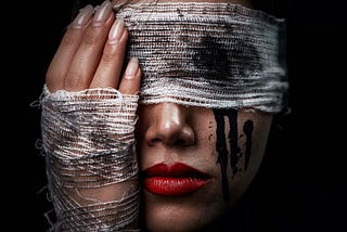 A lady with dark hair, a bandage stretched around her head covering her eyes, and wrapped around her hand that she is holding up to the side of her face.