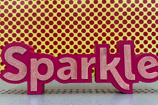 Pink sign that says “Sparkle” against a yellow backdrop with pink polkadots.
