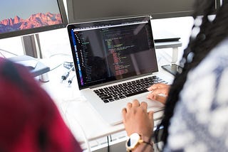 Dark skinned woman typing code on a laptop. We can also see the shoulder of someone else just outside of the frame of the photo.