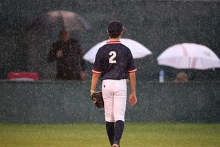 A 12-year-old baseball player stands alone on the infield grass in the rain.