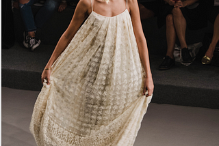 a woman on a runway wearing a long lacy spaghetti strap dress with the legs of the audience in the background. below the main image is a label that says “a woman in a dress holding a surfboard”