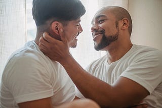 Two men wearing white tee-shirts embrace and lean in for a kiss.